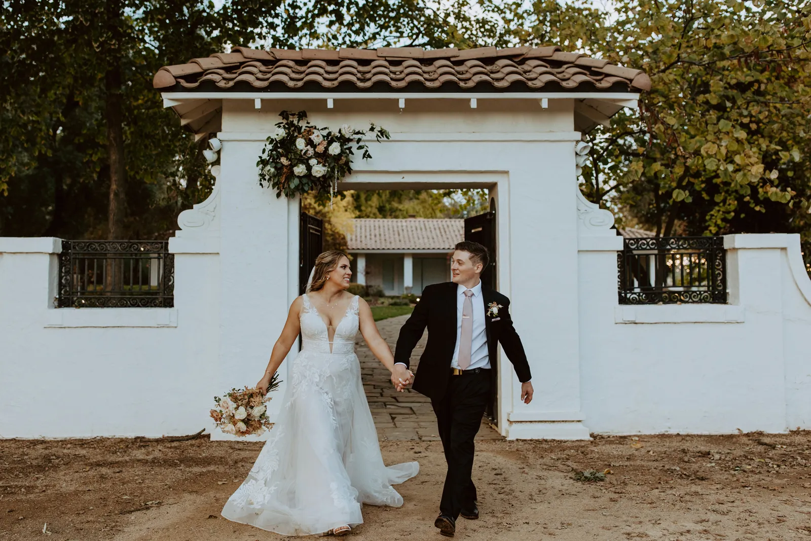 Bride and groom holding hands and walking out from underneath an archway.