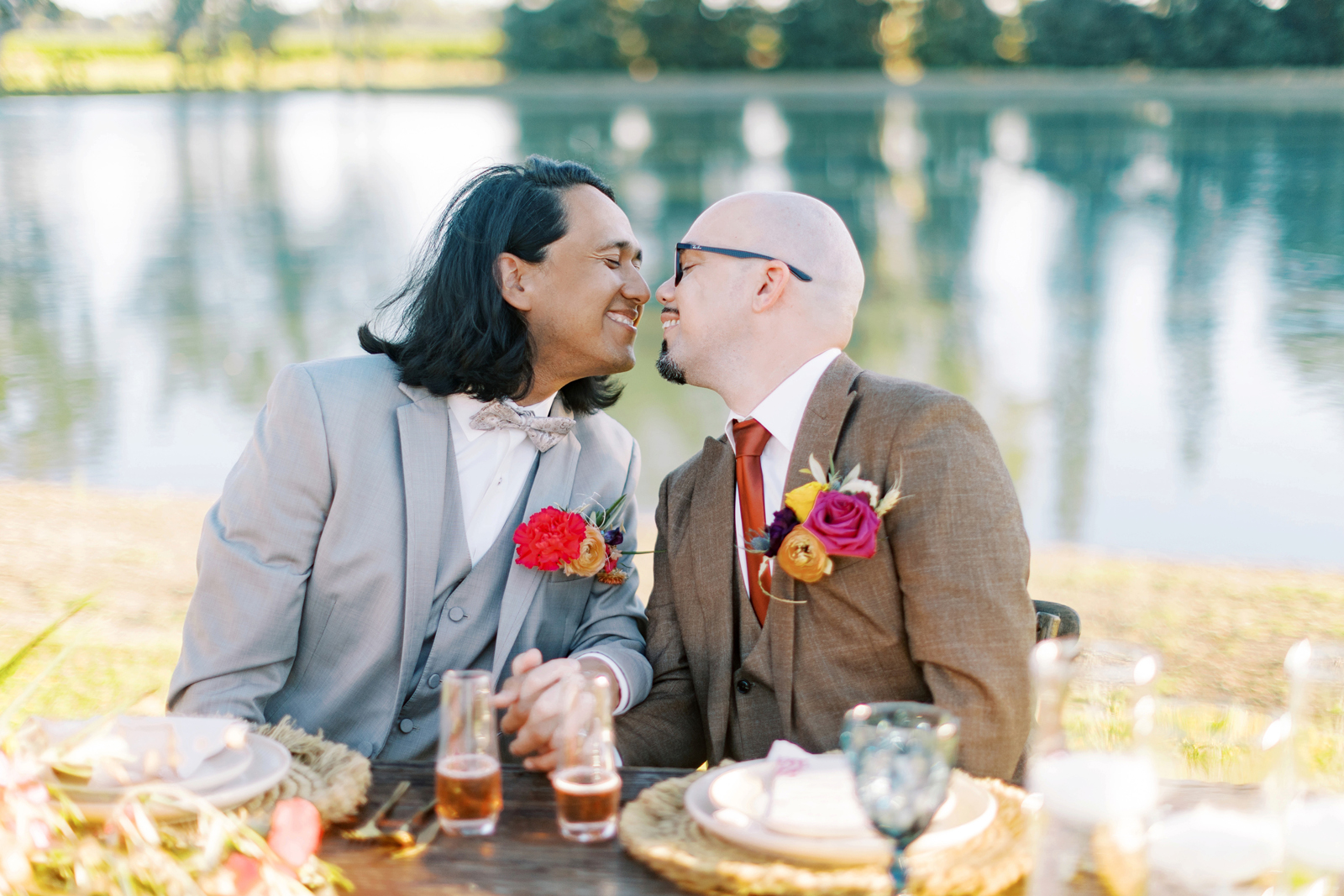 Grooms about to kiss at a table in front of a lake.