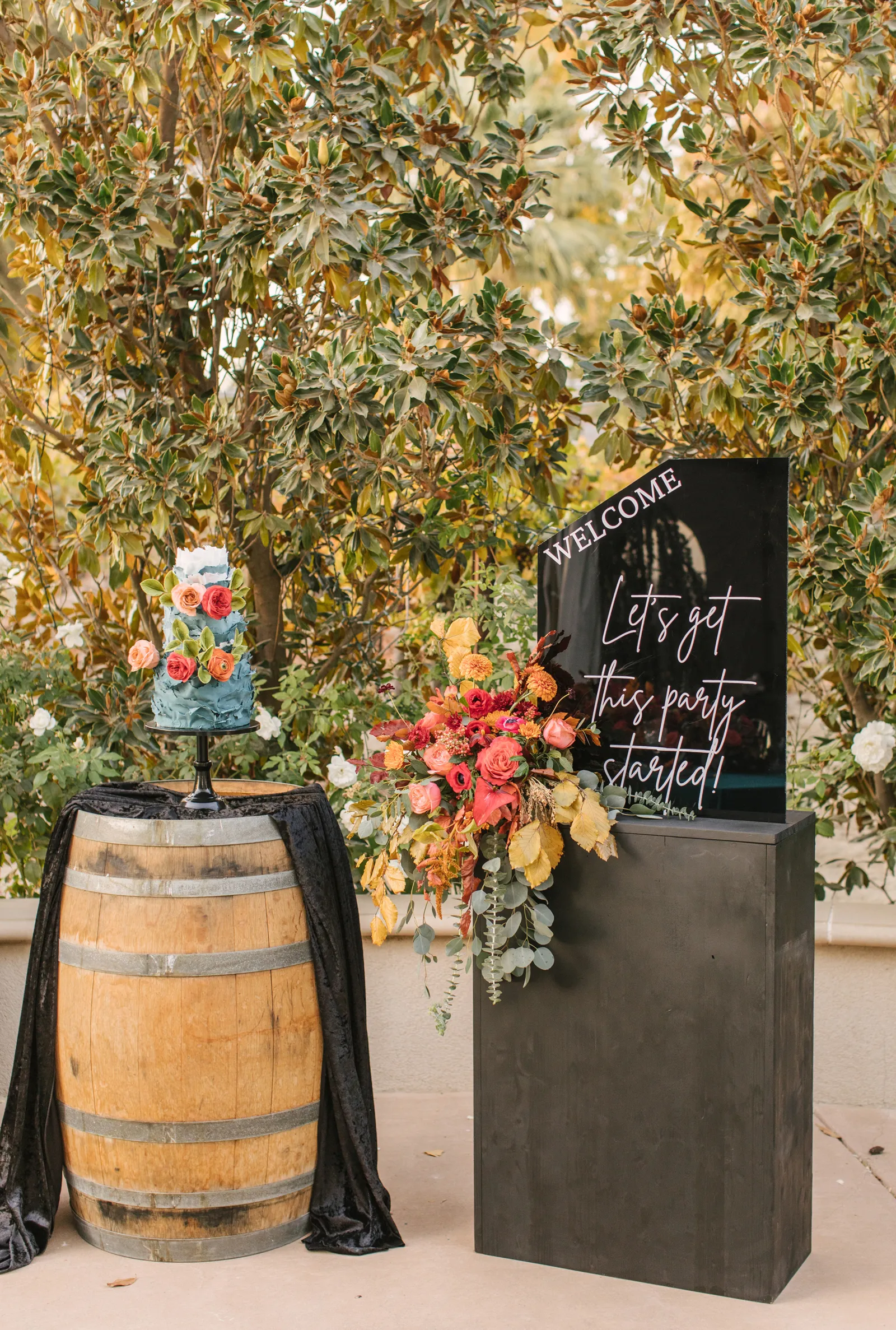 Barrel with flowers and a blue wedding cake and a black slab with words on it 