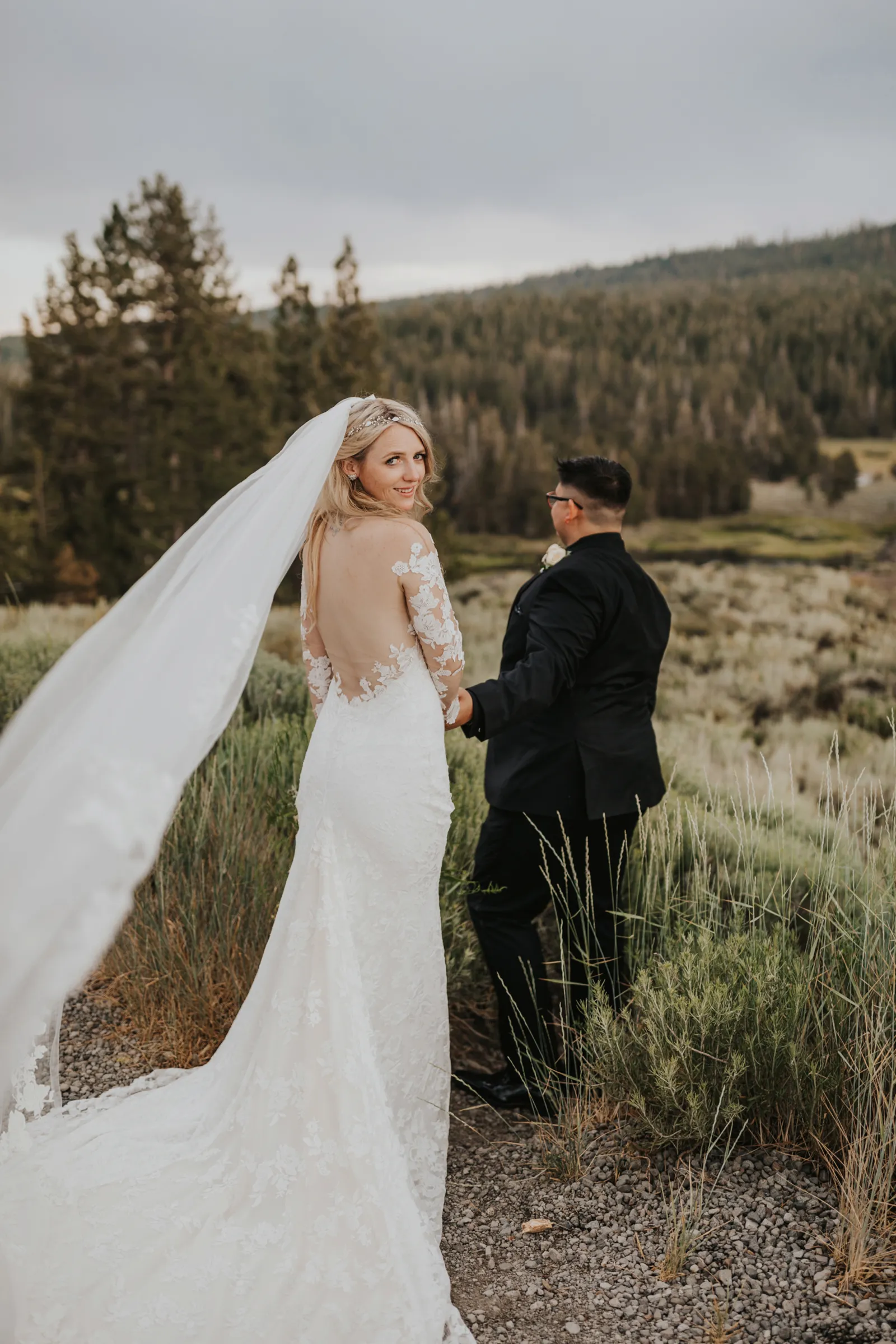 Bride looking over her shoulder as her groom leads her through the woods.