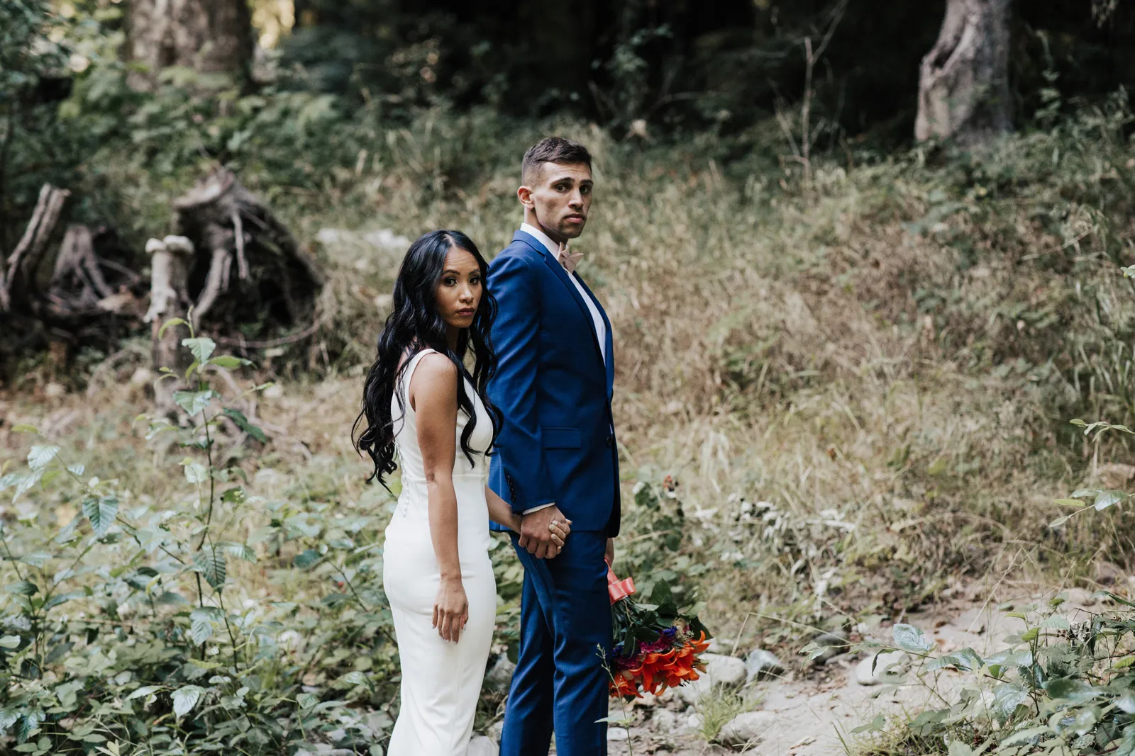 Bride and groom holding hands in the woods and looking over their shoulder at the camera.