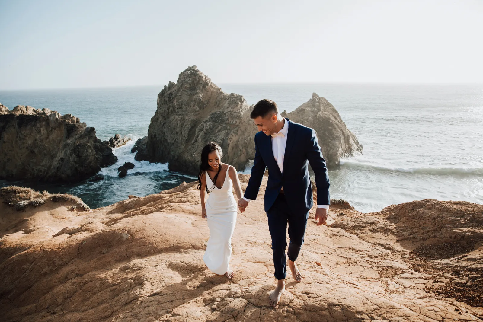 Bride and groom walking up a cliffside with the ocean behind them.