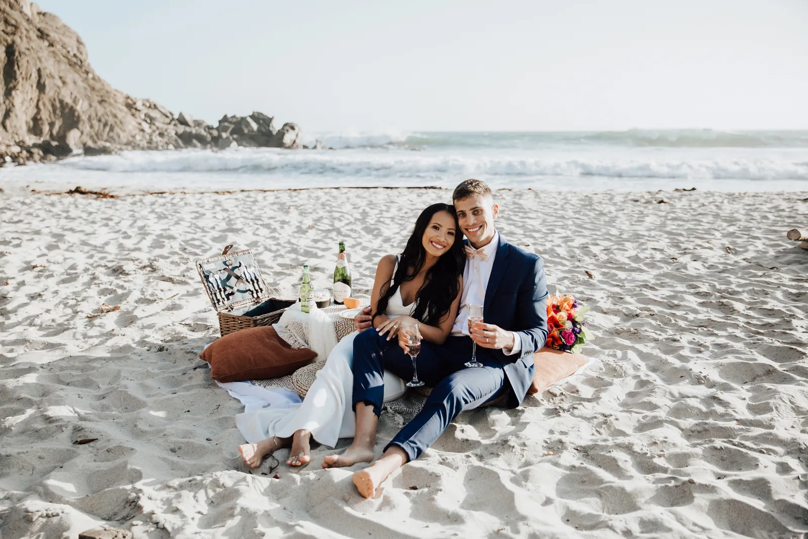 Bride and groom sitting by their beach picnic and smiling.