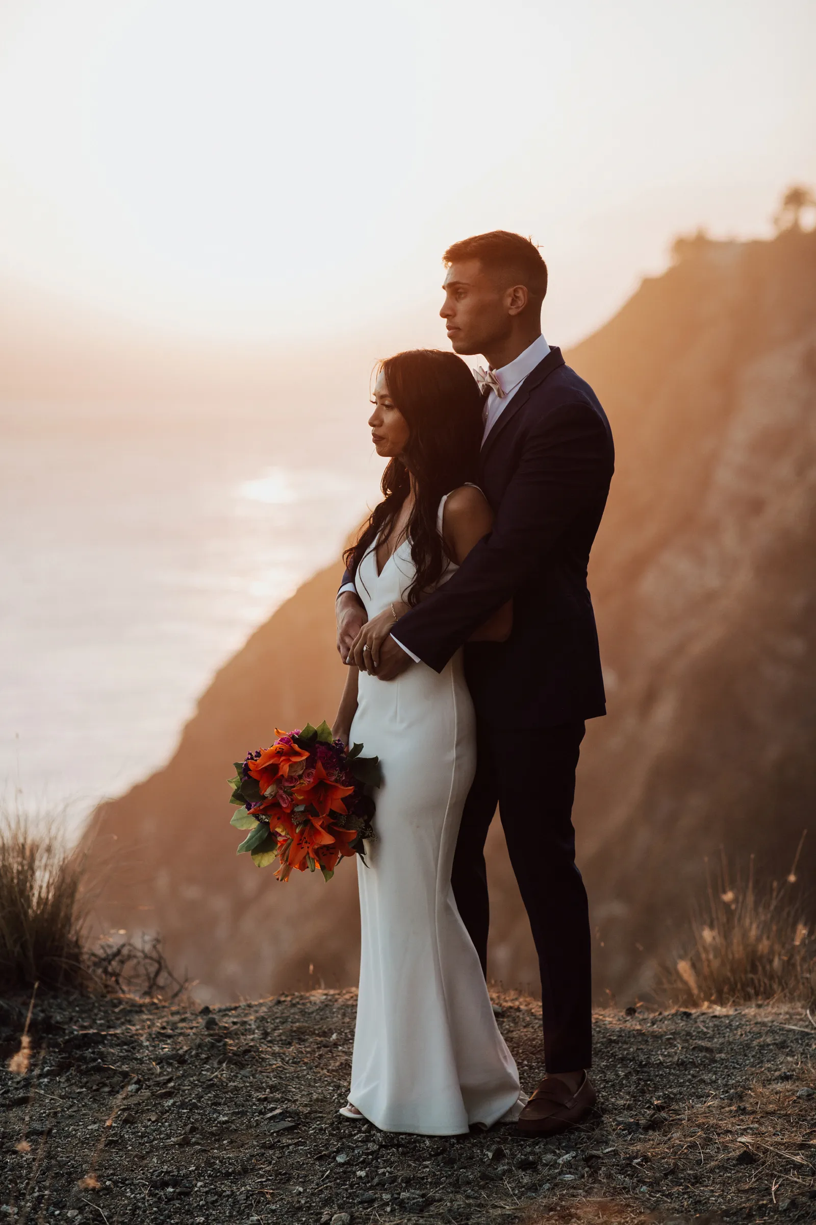 Groom holding his bride on a cliffside with the sunset behind them.