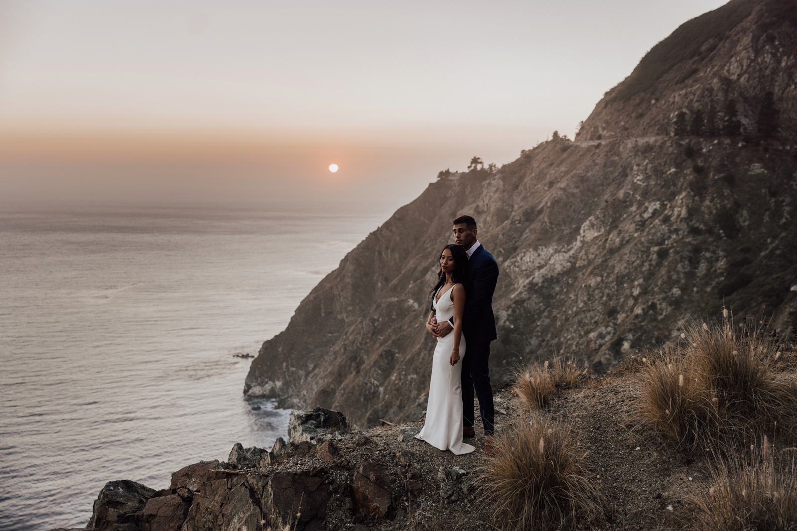 Groom and bride standing on a cliffside overlooking the ocean.