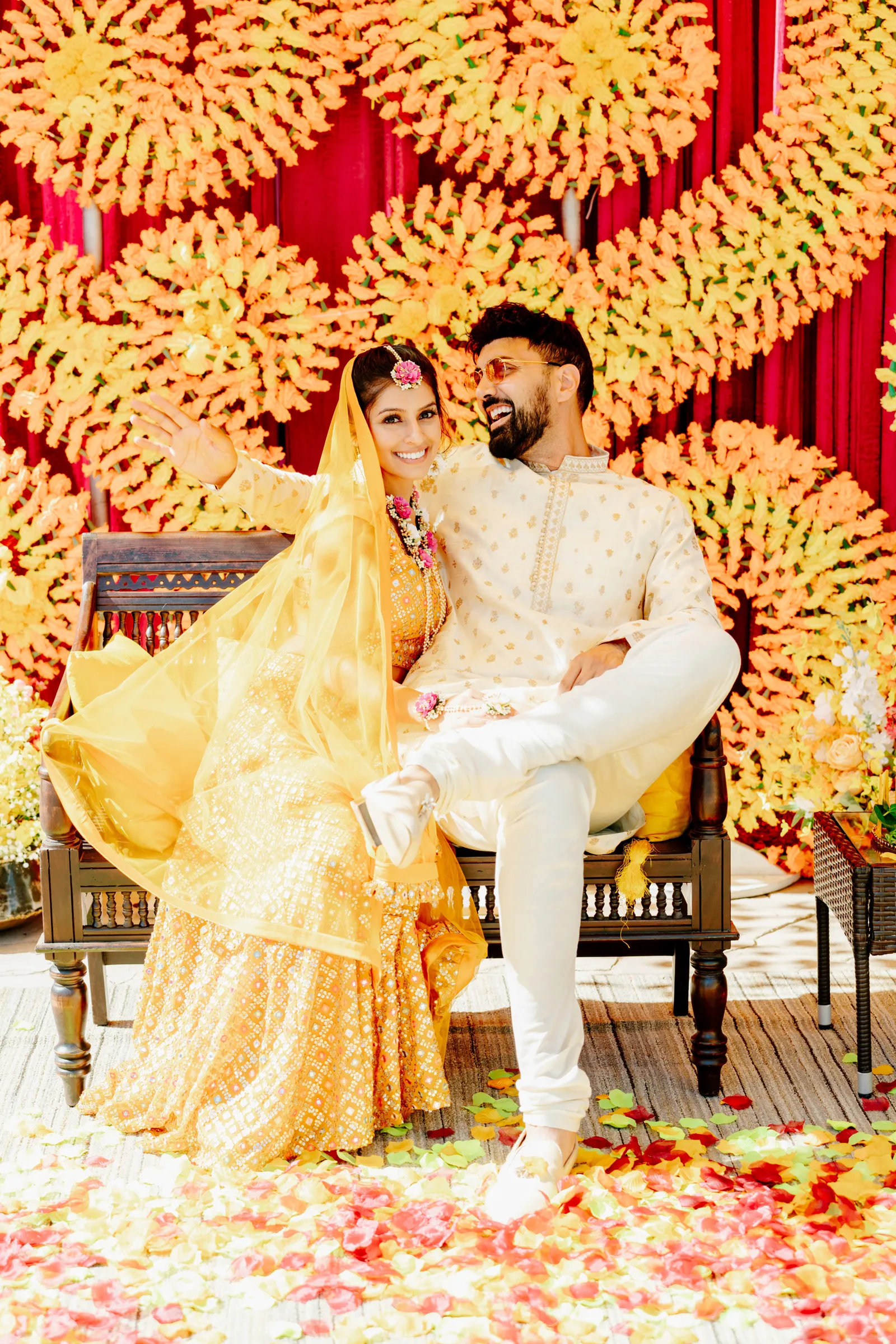 Bride and groom smiling together on a bench in their traditional, Indian wedding attire.