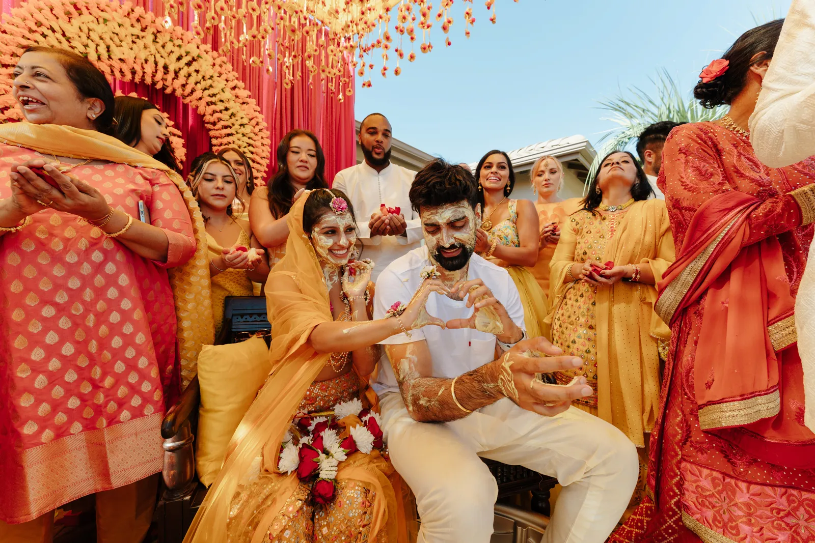 Bride and couple surrounded by friends and family and smiling, making a heart with their hands.