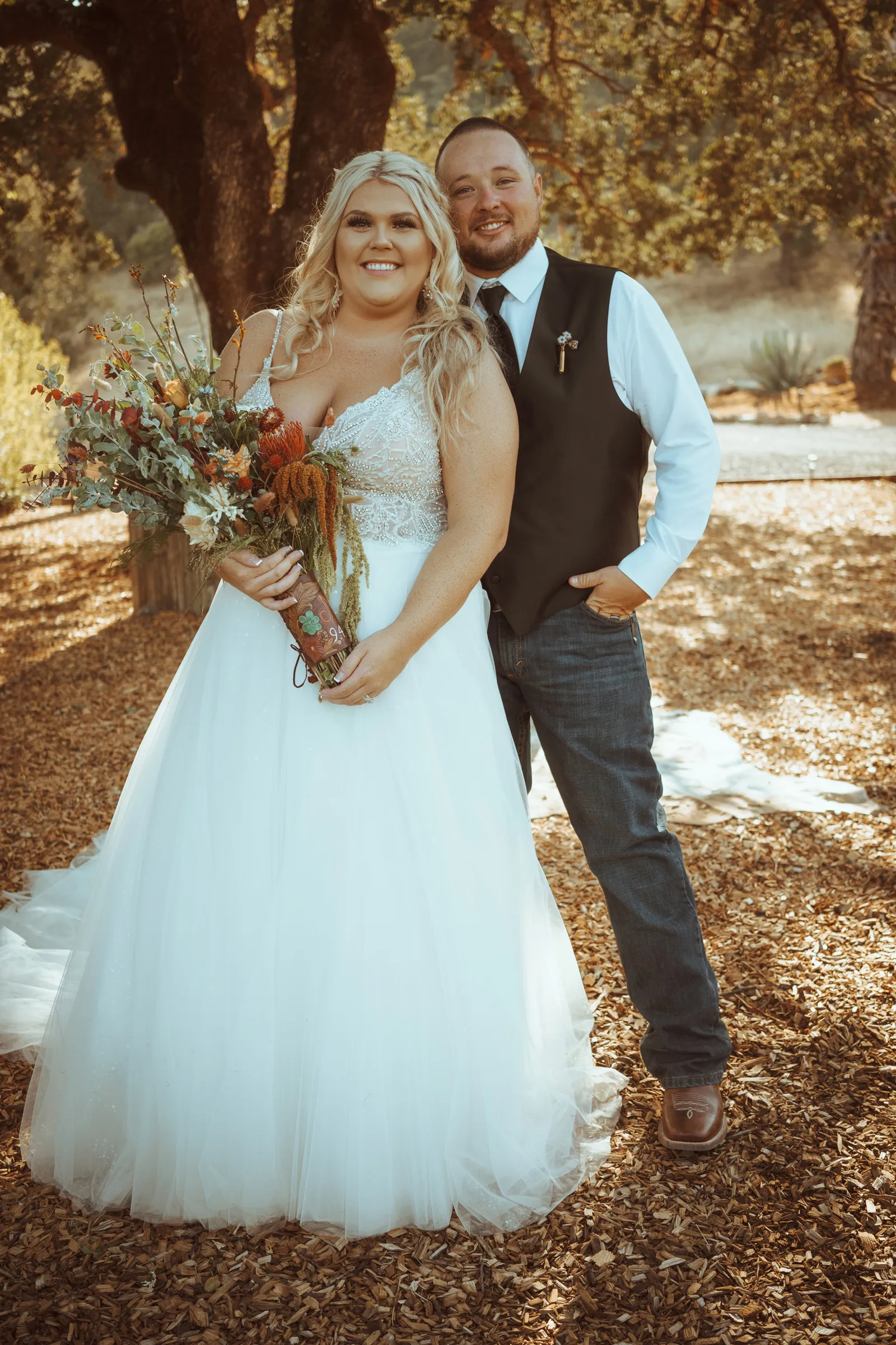 Bride and groom smiling outside by trees, posing for wedding photos.