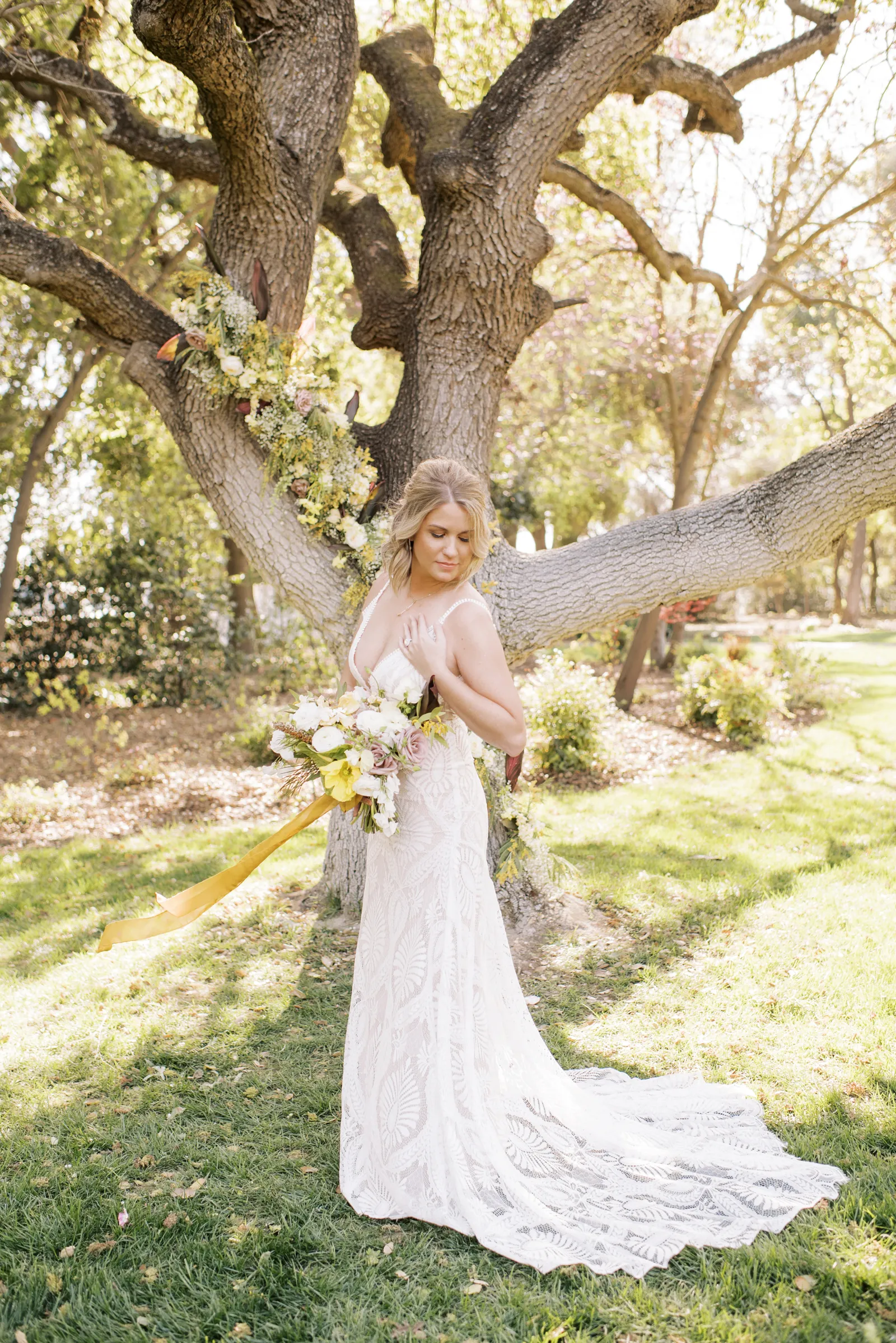 Bride smiling next to a tree and looking down at her bouquet.