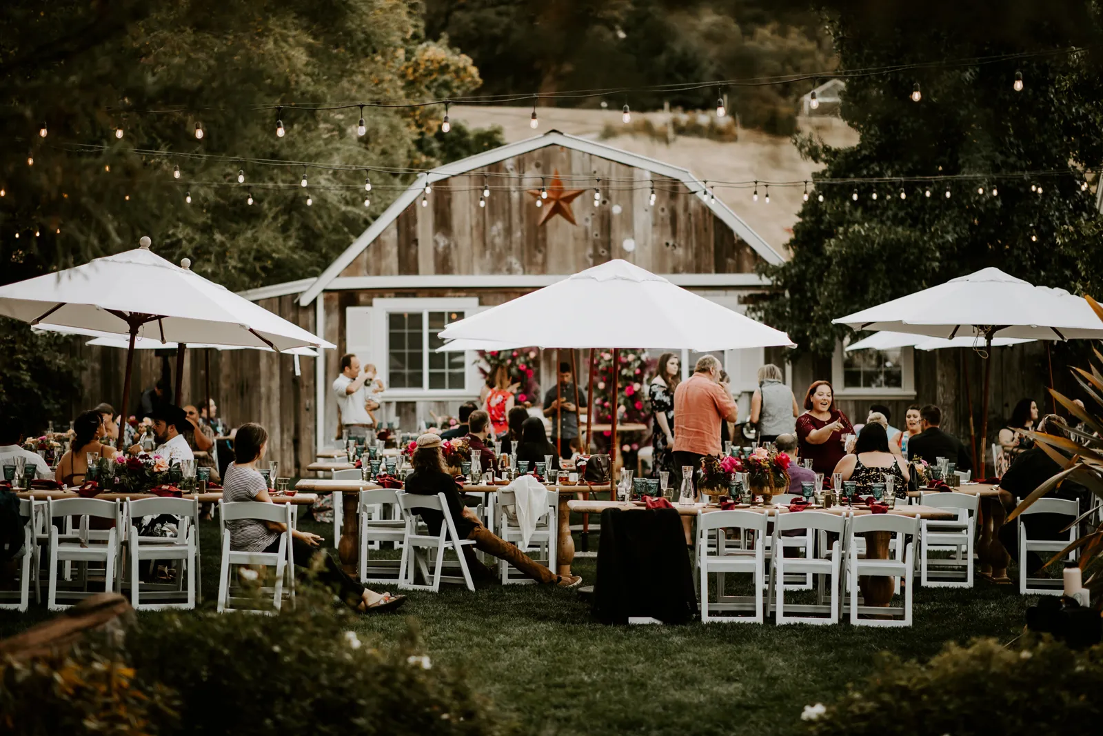 Photo of an outdoor venue with multiple chairs, tables and umbrellas next to a large barn.