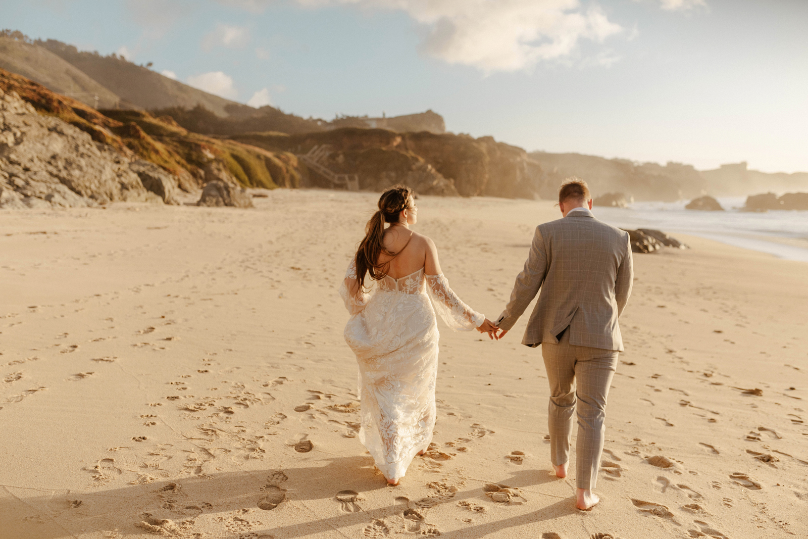 Bride groom holding hands with their backs to us, walking on the beach.