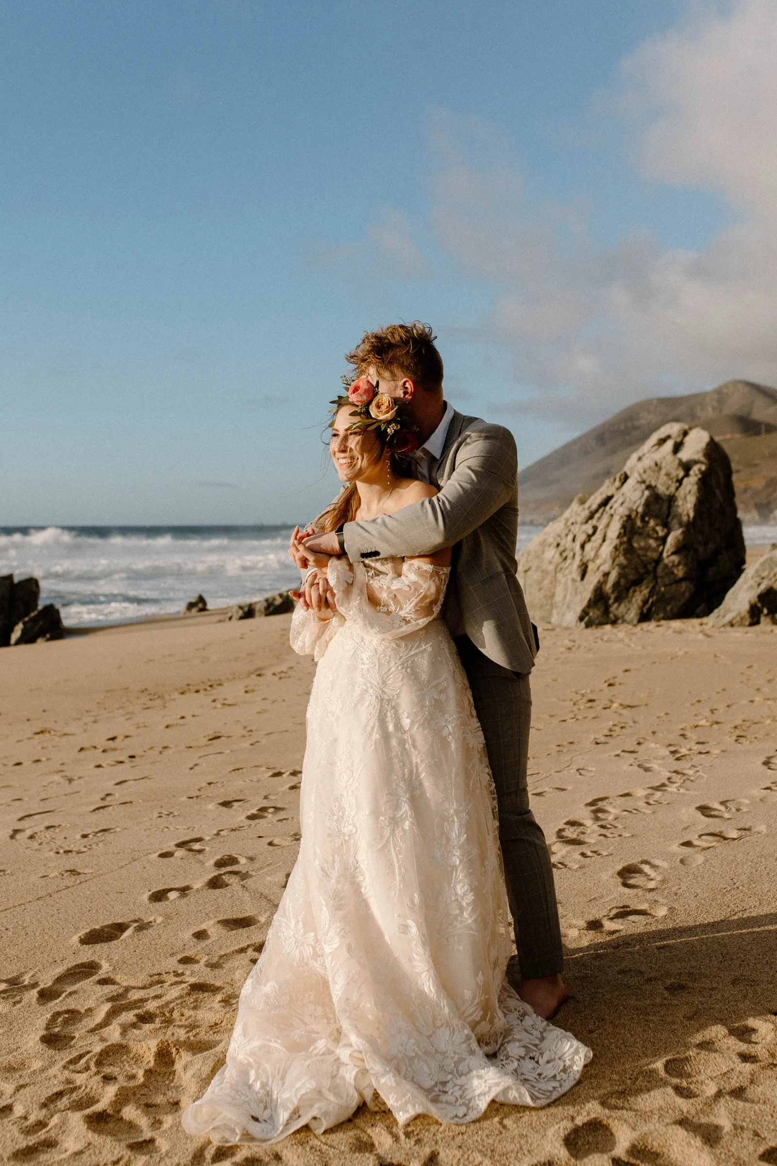 Groom hugging bride from behind on the beach.