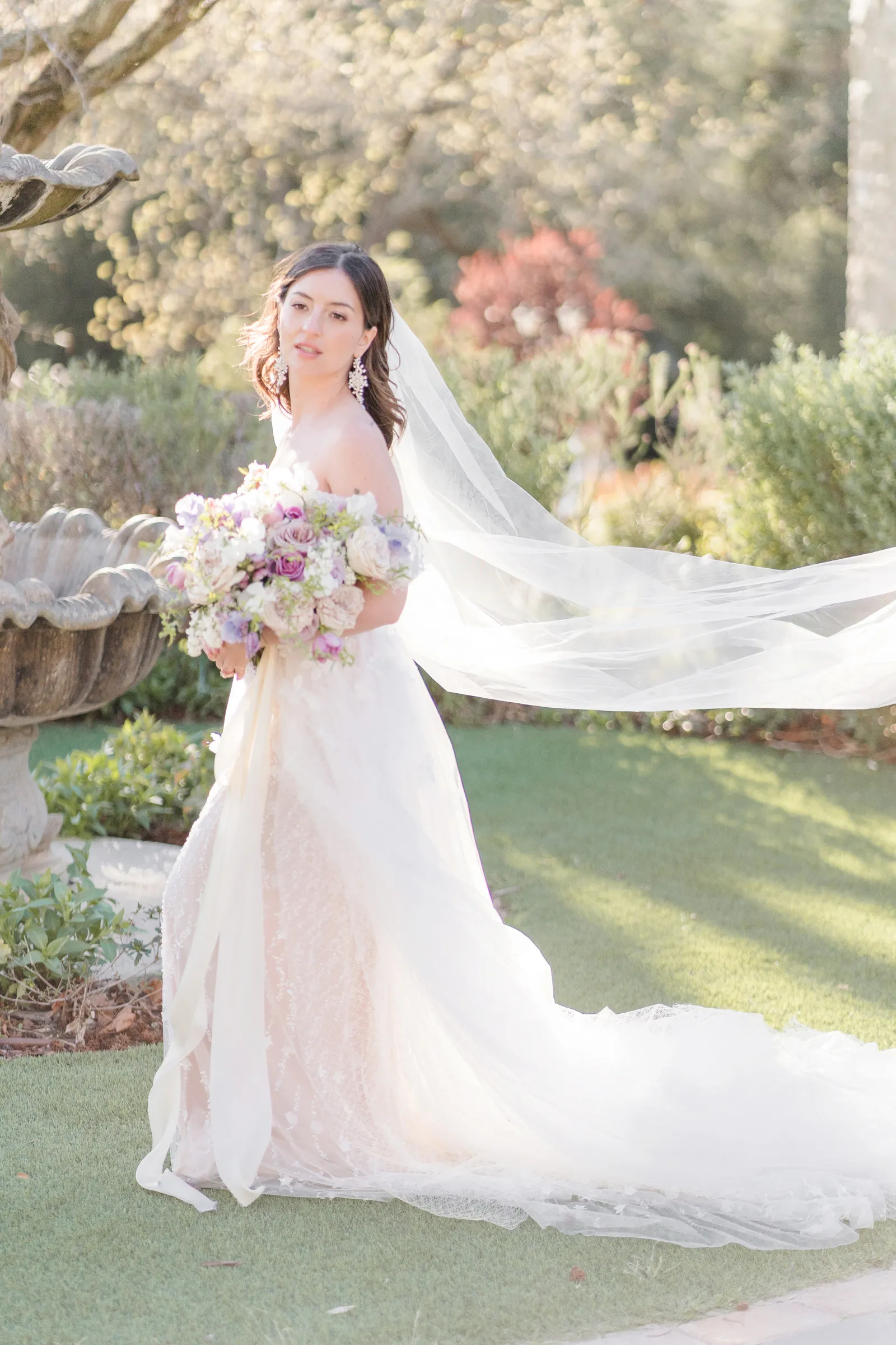 Picture of bride in her gown by a fountain, holding flowers.