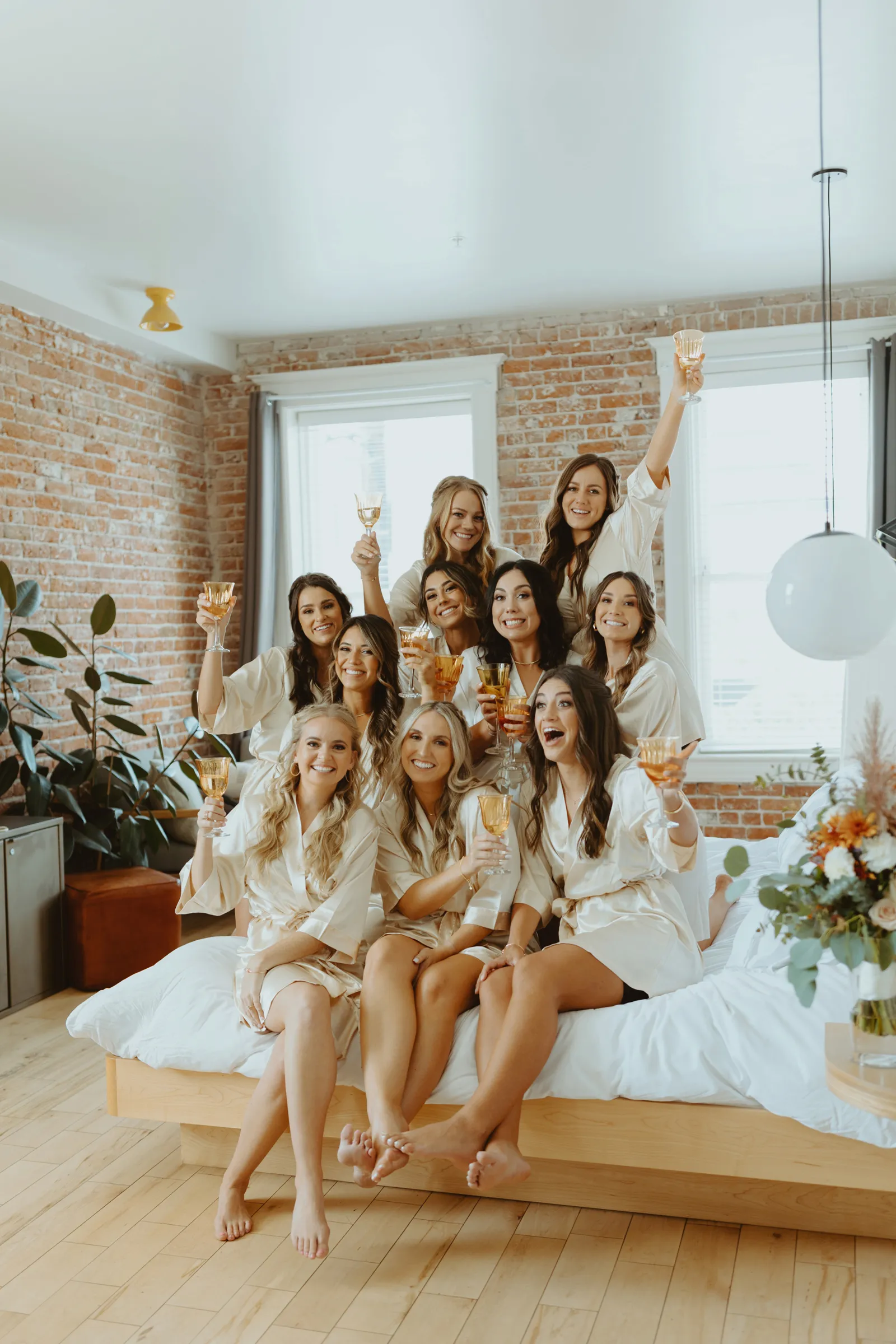 Bride and bridesmaids sitting on the bed together and holding up their glasses before getting ready.