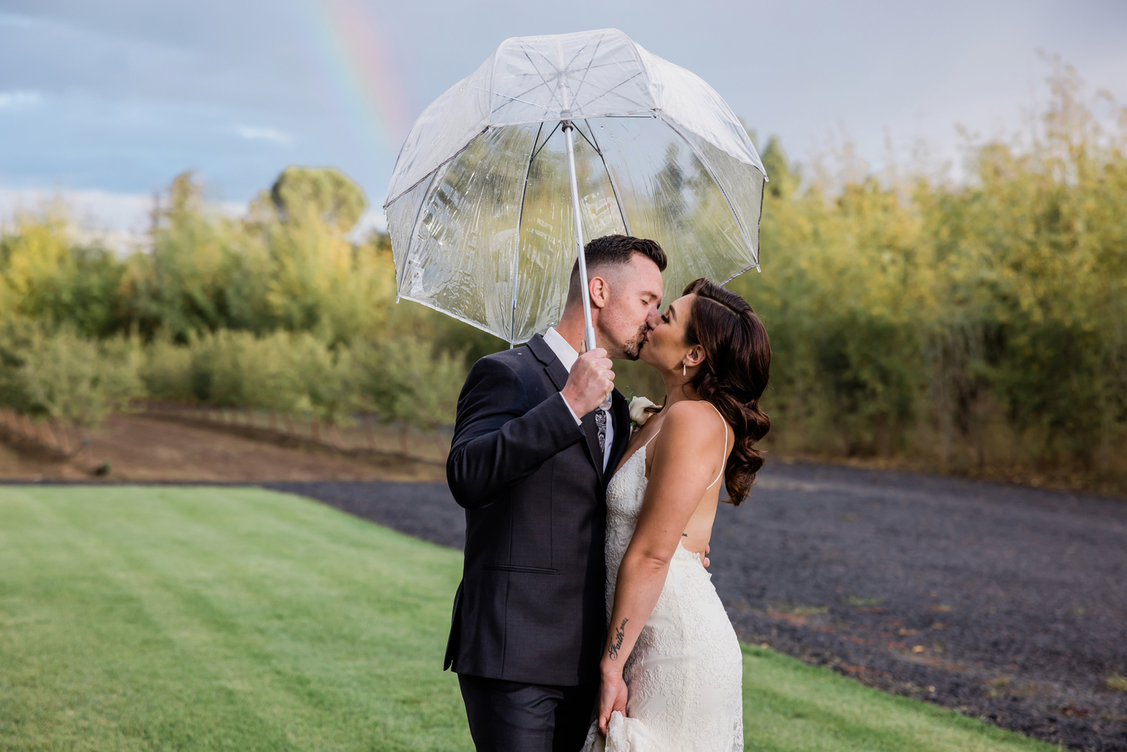 Groom holding an umbrella as he kisses his bride with a rainbow forming behind them.