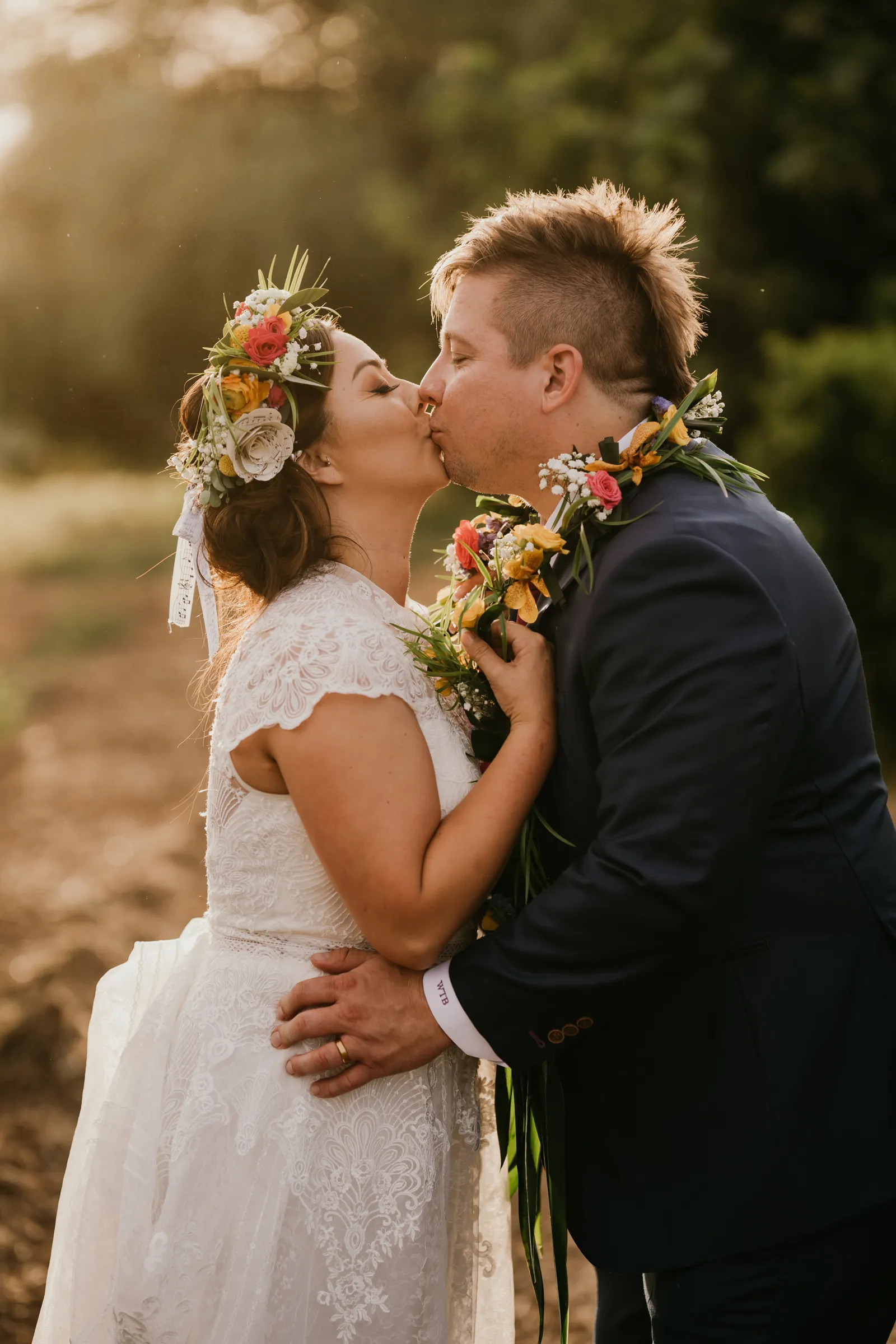 Bride and groom taking sunset photos, kissing with floral crowns and necklaces on.