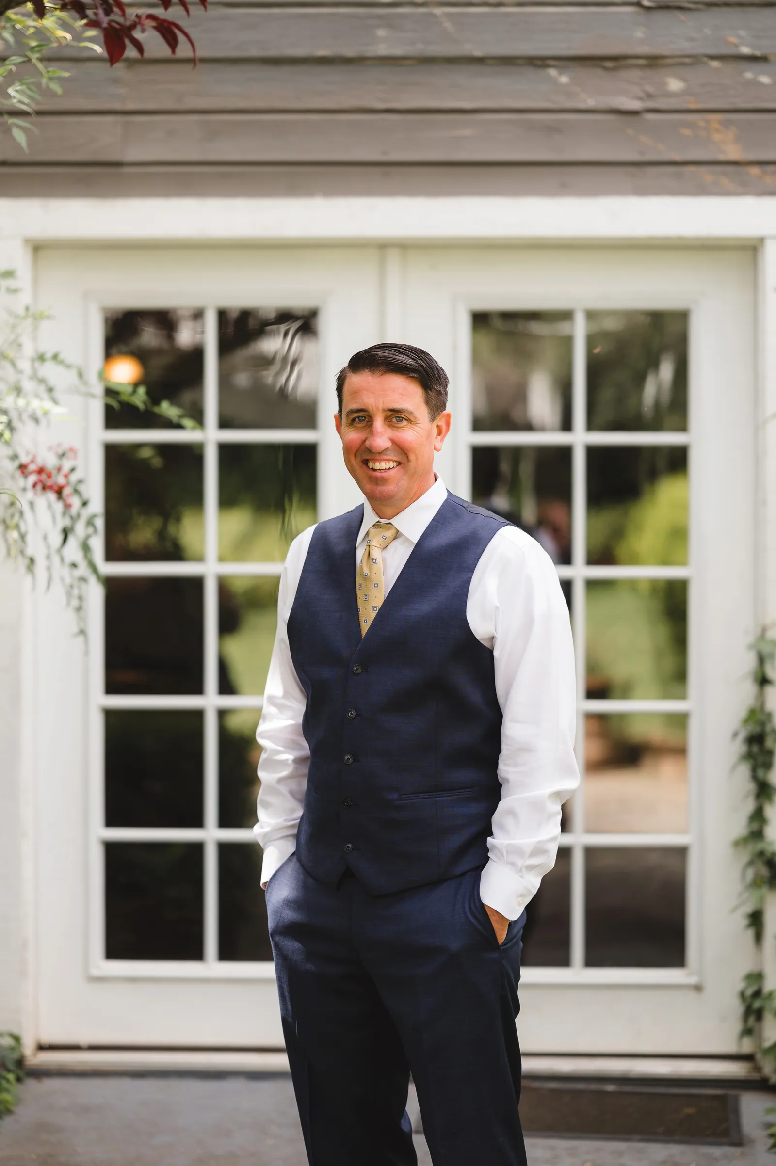 Groom smiling outside, with his hands in his pockets.