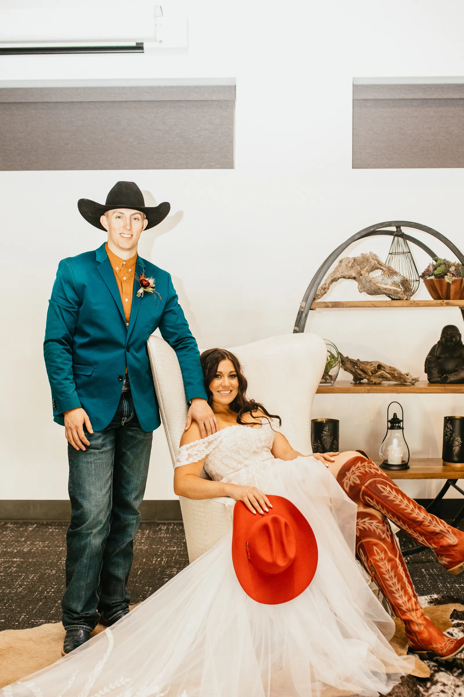 Bride sitting on a chair, holding a red cowgirl hat as her groom stands next to her.