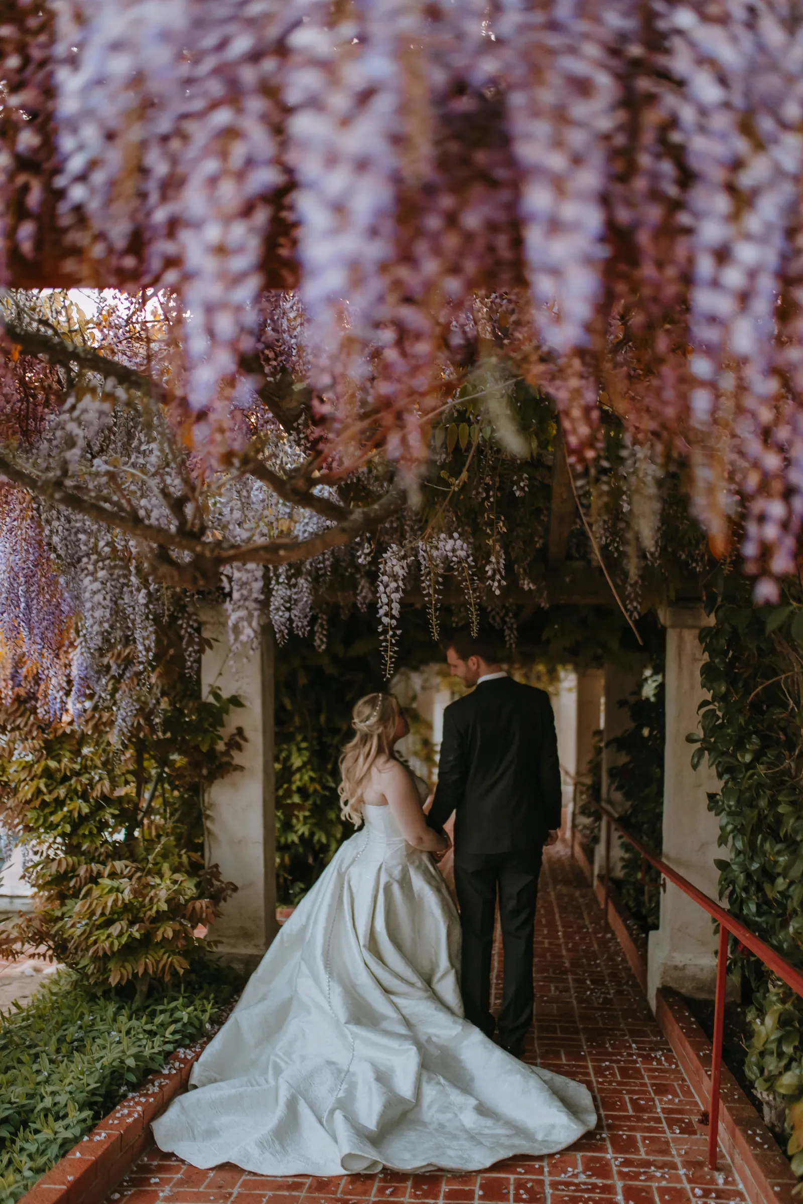 Newlyweds taking wedding photos under a canopy of pink and purple flowers.